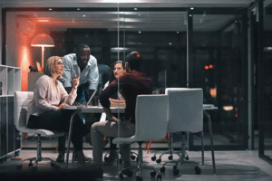 dois homens e duas mulheres em uma mesa de reunião em ambiente envidraçado. um dos homens está de pé explicando algo.
